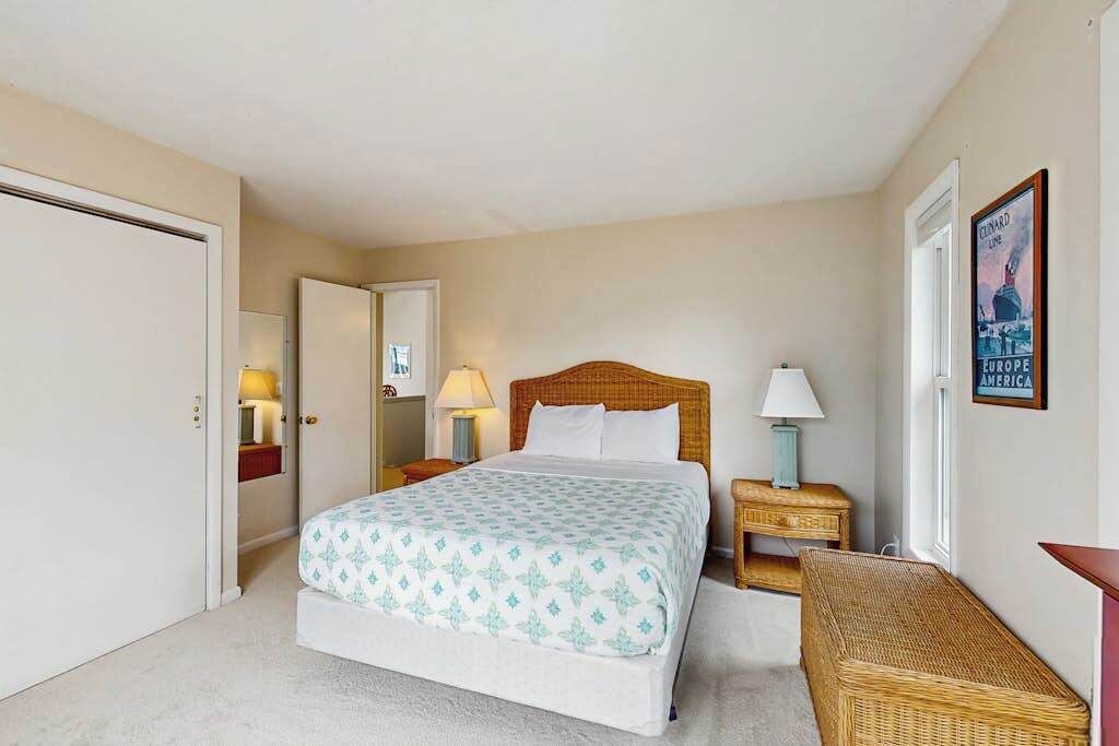 A bedroom with a bed, bedside tables, a lamp, a wicker bench, a door, and a poster on the wall.