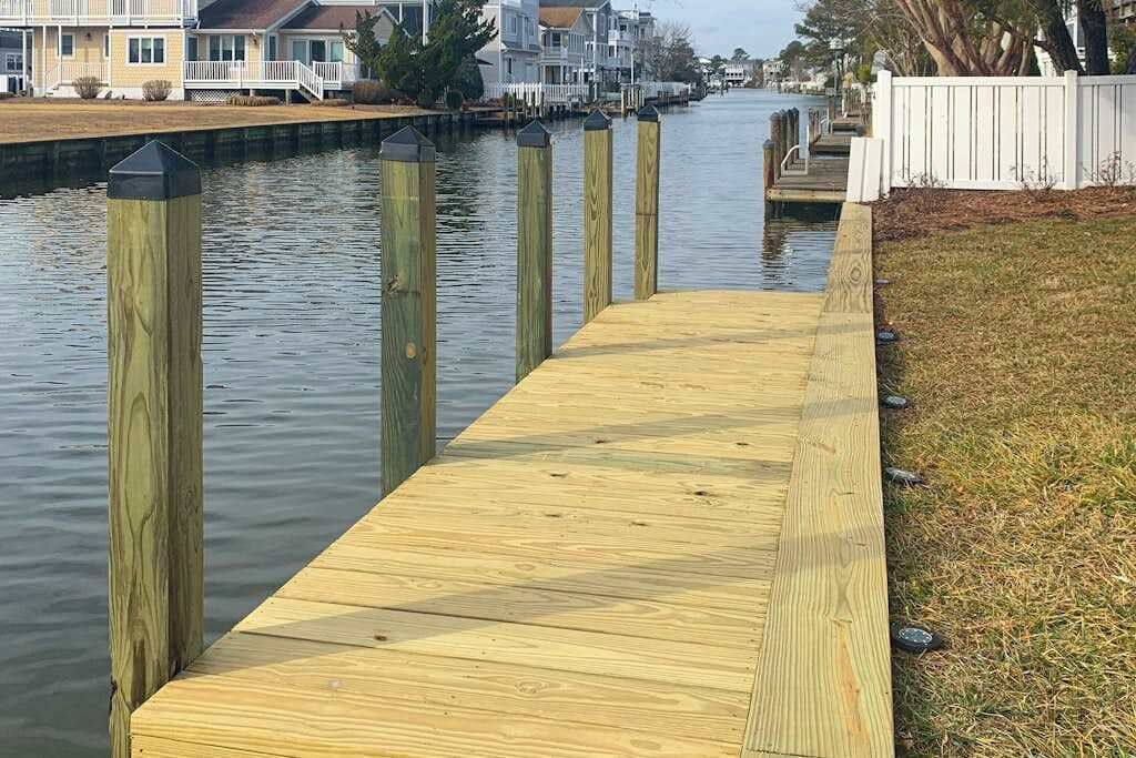A wooden dock extending into calm water, surrounded by a grassy area and a white fence.