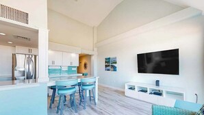 Una cocina moderna con taburetes de bar, zona de comedor, sala de estar con sofá y televisor montado.