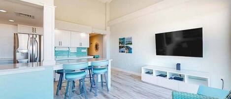 A modern kitchen with bar stools, a dining area, a living room with a sofa, and a mounted TV.