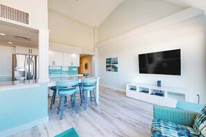 Eine moderne Küche mit Barhocker, einem Essbereich, einem Wohnzimmer mit Sofa und einem an der Wand befestigten Fernseher.