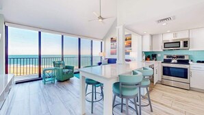 Una cocina moderna con una isla central, un comedor con sillas y vistas a la playa a través de amplios ventanales.