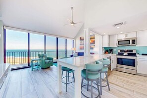 A modern kitchen with a central island, a dining area with chairs, and a view of the beach through large windows. - Sea Colony Ocean Penthouse condo with balcony, beach access, pools, & hot tub (Bethany Beach)