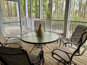 A covered outdoor seating area with a round table and chairs, a decorative vase, and a view of a wooded area through large windows. - Bear Trap Dunes first floor condo w/community pool, tennis court & on site golf (Ocean View)