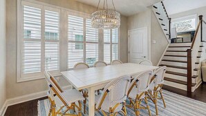 Uma sala de jantar com uma mesa branca e cadeiras de vime, um candelabro e uma escada.