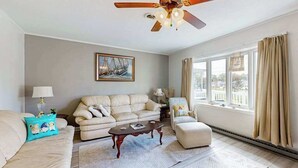 Un salon avec un ventilateur de plafond, un canapé, un fauteuil, une table basse et une fenêtre avec des rideaux.
