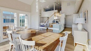 A spacious dining area with a large wooden table and white chairs, a staircase leading to an upper level, and a view of the outside. - Spacious pet friendly waterfront house w/deck, patio, balcony & gas grill (Bethany Beach)