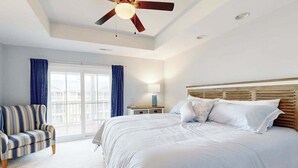 Een slaapkamer met een groot bed, een plafondventilator, een nachtkastje, een lamp, een raam met gordijnen en een gestreepte stoel.
