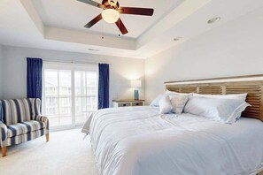 A bedroom with a large bed, a ceiling fan, a nightstand, a lamp, a window with curtains, and a striped chair. - Sandy Cove Townhouse w/ Water Views & Shared Pool, Private W/D, Free Wifi (Ocean View)