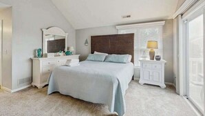 A bedroom with a bed, dresser, nightstand, and a large window with blinds. - Bay view condo with screened porch, pickleball, on-site golf and shared pool (Ocean View)
