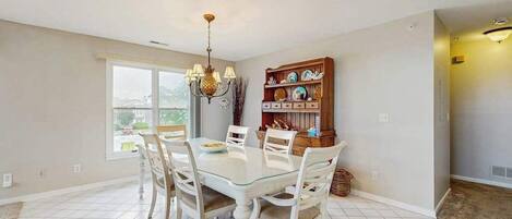 A dining area with a glass table, white chairs, a chandelier, and a bookshelf.