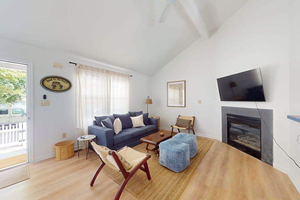 Uma sala de estar com sofá, banquinho, poltrona, lareira, televisão e uma janela com cortinas.