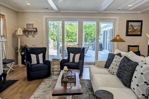 A living room with a sofa, two armchairs, a coffee table, and a view through glass doors to an outdoor deck. - Town of Bethany Beach house w/private pool, hot tub, games, deck, grill (Bethany Beach)