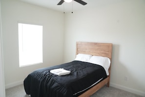 Een slaapkamer met een enkel bed, een plafondventilator, een raam en een effen muur.