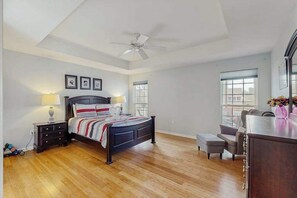 Quarto com cama, mesas de cabeceira, escrivaninha, cadeira, ventilador de teto e janelas amplas.