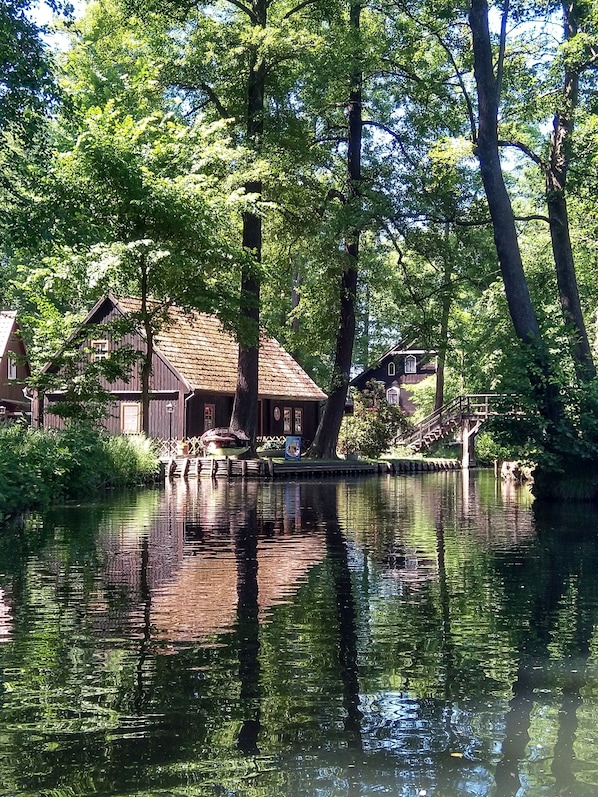 Ein hölzernes Haus am Flussufer mit Spiegelung im Wasser, umgeben von Bäumen.