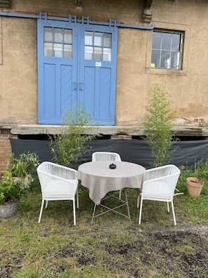 Ein kleiner Außenbereich mit Tisch und zwei Stühlen, einer blauen Tür und einem Fenster.