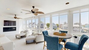 A spacious living room with a large window, a dining area with blue chairs, and a ceiling fan. - Keen-Wik on the Bay bay front home w/private pool & hot tub (Selbyville)