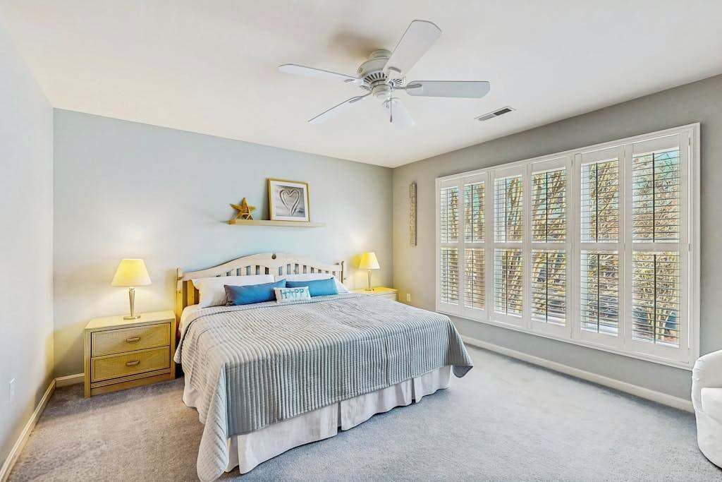 Ein Schlafzimmer mit Bett, Nachttischen, Deckenventilator und einem großen Fenster mit Jalousien.