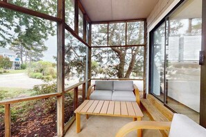 Eine Holzterrasse mit einer Couch und einem Tisch, großen Fenstern und Blick auf Bäume und eine Straße.