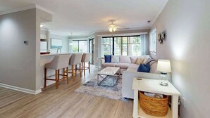 Un salon moderne comprenant un canapé, une table basse et des fauteuils, un coin cuisine avec une table à manger, et de grandes fenêtres laissant entrer la lumière naturelle.
