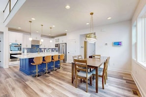 Una cocina moderna con una isla central, taburetes de bar, una mesa de comedor con sillas, iluminación colgante y un televisor montado en la pared.