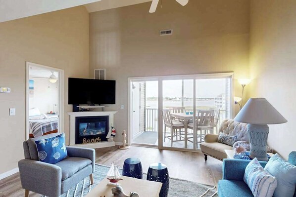 A living room with a fireplace, a television, and a balcony with outdoor furniture. - Bethany Bay 3rd floor condo w/pool, hot tub, tennis court, on-site golf (Ocean View)