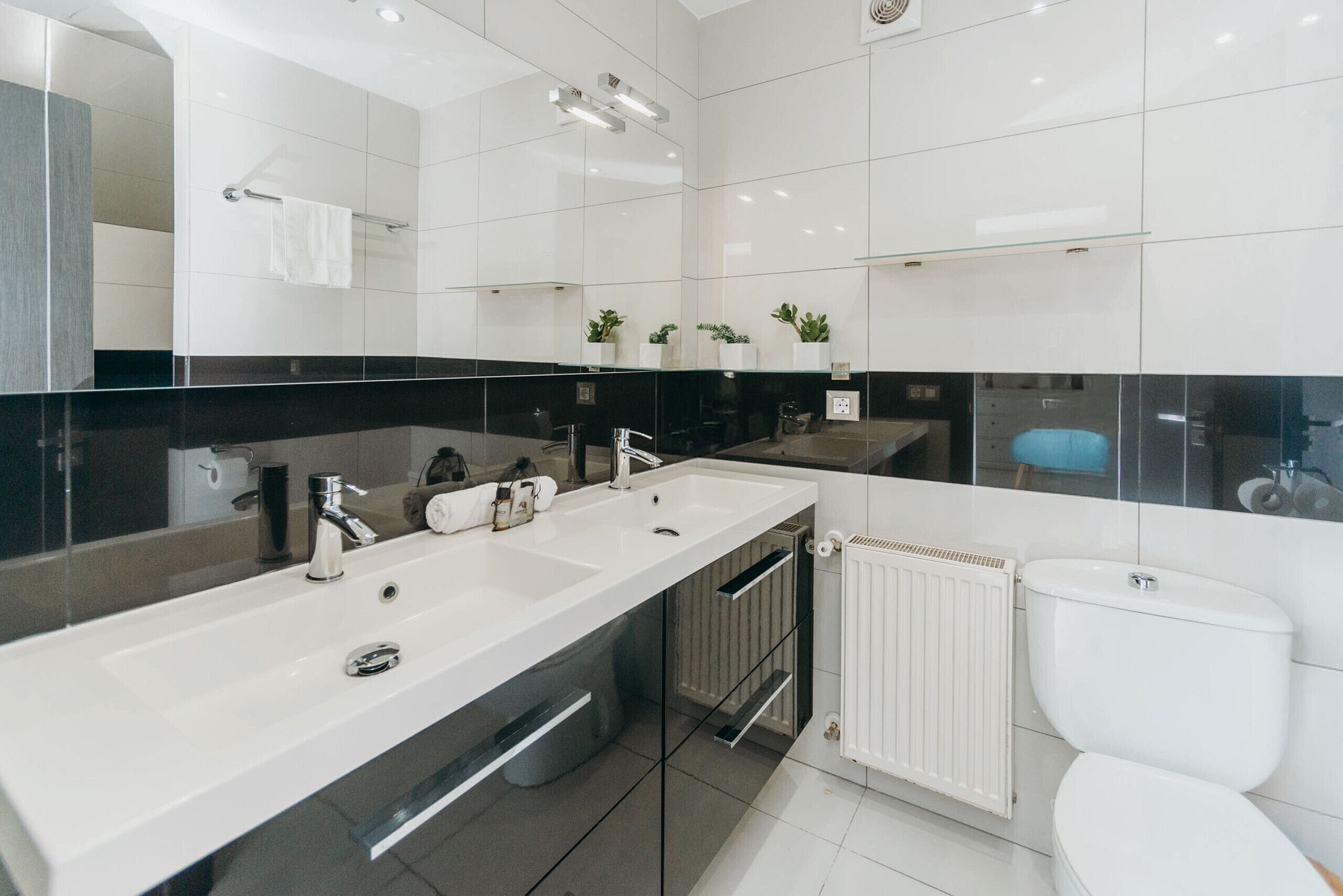 A modern bathroom with a large mirror, two sinks, a toilet, and a radiator.