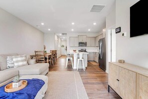 A modern living room with a sofa, dining area, kitchen, and a flat-screen TV mounted on the wall. - Millville By The Sea - 36692 Calm Water Dr | Townhouse in Millville (Millville)