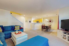 Ein Wohnzimmer mit einem blauen Sofa, einem weißen Couchtisch, einem Essbereich mit Tisch und Stühlen, einer Küche mit gelben Schränken und einem Fernseher auf einem Schrank.