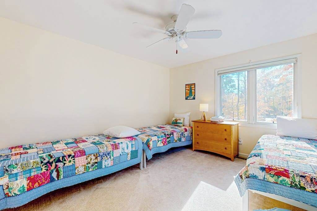 A bedroom with two single beds, a ceiling fan, a dresser, a window with a view, and a painting on the wall.