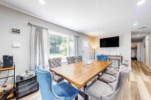 A modern dining area with a wooden table, upholstered chairs, a flat-screen TV, and a view of the outdoors. - Town of Millville - 32669 Cedar Drive | House in Millville (Millville)