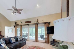 A living room with a leather sofa, a ceiling fan, a TV, and a large window with curtains. - Salt Pond house with golf course views, pool, tennis, fireplace, porch, deck, AC (Bethany Beach)