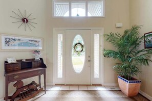 A foyer with a white door, a wooden console table, a large potted plant, a framed picture, and a starburst clock. - Salt Pond house with golf course views, pool, tennis, fireplace, porch, deck, AC (Bethany Beach)