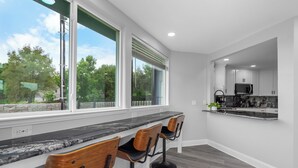 A modern kitchen with a marble countertop, wooden bar stools, and a view of trees through large windows. - The Lone Star Lakehouse at Eagle Mountain Lake (Azle)