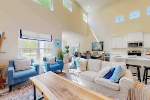 Ein geräumiges Wohnzimmer mit einem großen Holztisch, zwei blauen Sofas und einem weißen Sofa mit blauen Kissen. Im Hintergrund ist eine Küche mit weißen Schränken und einer Mikrowelle zu sehen.