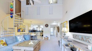 A spacious living area with a sofa, coffee table, TV, and a staircase with colorful decorations. - Sea Colony home with loft, sauna, pool, tennis court, hot tub, & balcony (Bethany Beach)