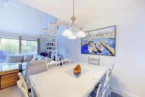 Ett modernt vardagsrum med en vit soffa, ett matbord med en ensam fralla, en vit steghylla och en stor tavla som föreställer en hamnmiljö.