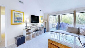 A modern living room with a TV, a sofa, a glass coffee table, and a balcony with a view. - Sea Colony home with loft, sauna, pool, tennis court, hot tub, & balcony (Bethany Beach)