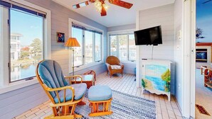 Una stanza luminosa con ventilatore a soffitto, televisore, un quadro e vista su un corpo d'acqua attraverso ampie finestre.