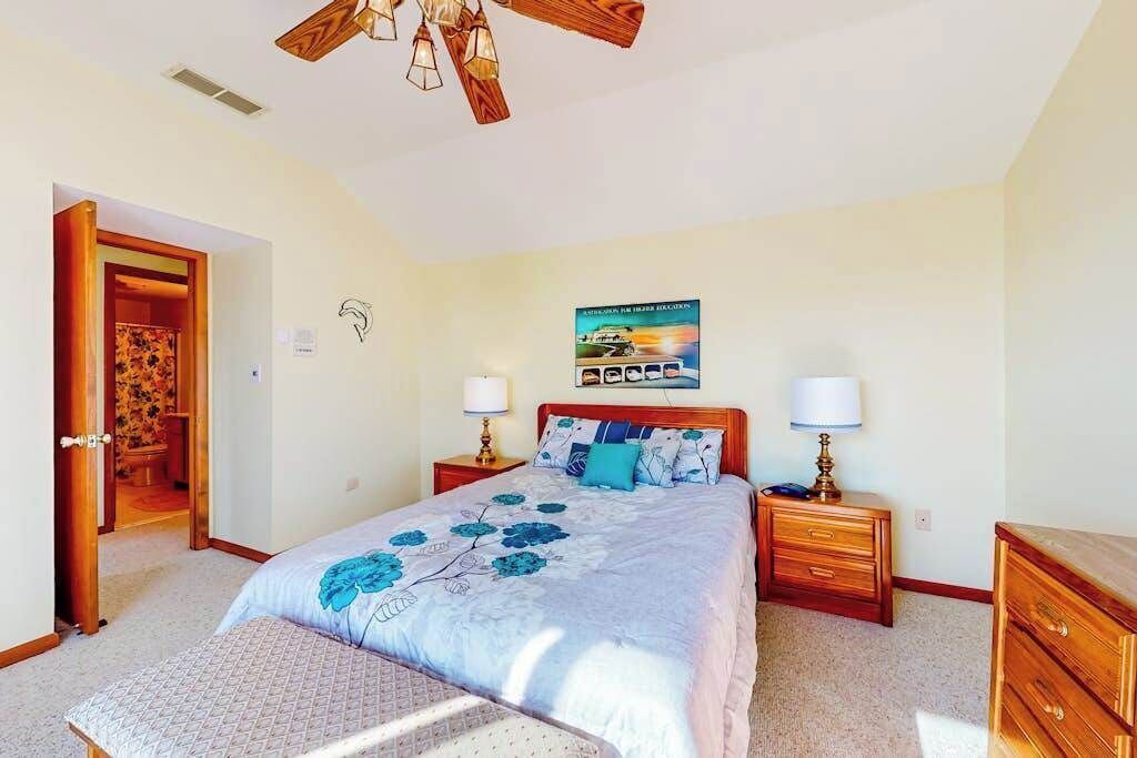 Une chambre à coucher comprenant un lit, des tables de chevet, une tête de lit en bois, un ventilateur de plafond et une porte donnant sur une autre pièce.
