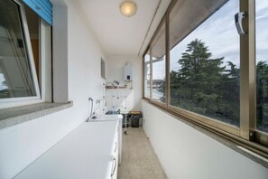 A narrow balcony with a washing machine, a small sink, and a window with a view of trees. - A comfortable and welcoming 60 square meters apartment, perfect for four people, located on the fourth floor of a period building without an elevator.The property is located in a quiet area, allowing our guests to easily and quickly reach the center of U (Udine)