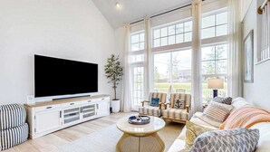 Un salon avec un téléviseur, un canapé, des fauteuils et de grandes fenêtres.