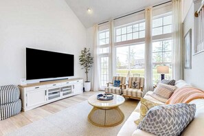 A living room with a TV, sofa, chairs, and large windows. - Marlin Chase home with resort amenities, bikes, grill, EV charger - dog-friendly (Ocean View)