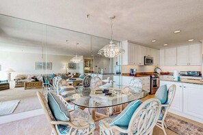 Eine moderne Küche mit weißen Schränken, ein Essbereich mit einem Glastisch und Korbstühlen sowie ein Wohnzimmer mit einer Couch und einem Teppich.