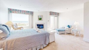 A bedroom with a bed, two chairs, a small table, a fireplace, and large windows. - Kings Grant Beachfront Townhouse w/Ocean Views, Shared Pool, Free WiFi, Private (Fenwick Island)