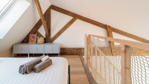 Een slaapkamer met een schuine balkenplafond, een bed met opgerolde handdoeken, een klein dressoir en een raam.