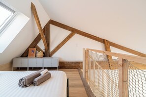 A bedroom with a sloped ceiling, wooden beams, a bed with rolled towels, a small dresser, and a window.