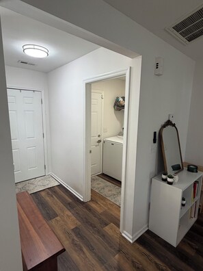 Un couloir avec un banc en bois, une porte blanche, un meuble blanc avec un miroir et un petit réfrigérateur.