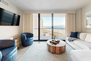 A modern living room with a large TV, a round coffee table, a sofa, and a chair. - Sea Colony Ocean renovated beachfront condo with balcony, AC, pools, & tennis (Bethany Beach)